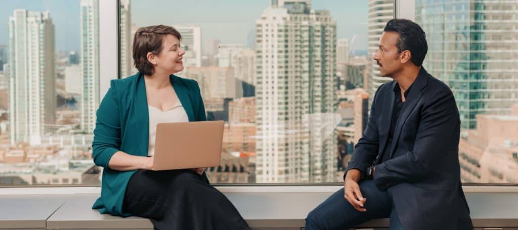 A woman holding a laptop and a man in business attire have a professional conversation in a modern office with a city skyline in the background. The setting suggests a collaborative meeting, symbolizing the process of hiring or consulting with digital accessibility experts.