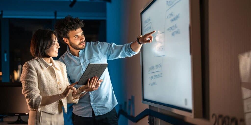 Two colleagues review AI transformation plans on a digital whiteboard, discussing roles, workflows, and opportunities to apply AI across the business. This illustrates the human collaboration and strategic work necessary to become an AI-first organization.